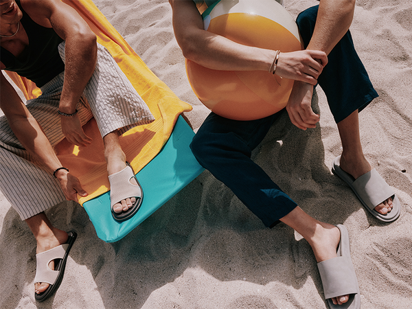 Two men are on the beach. One sits on a blue chair with a yellow towel, wearing striped pants and beige-and-black slide sandals. The other sits on the sand holding a beach ball, wearing navy pants and beige slide sandals.