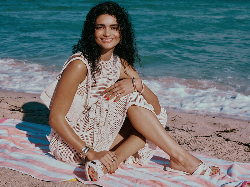 A woman sitting on a pink-and-white striped beach towel, wearing a crochet dress and accessorized with a white handbag and white flat sandals.
